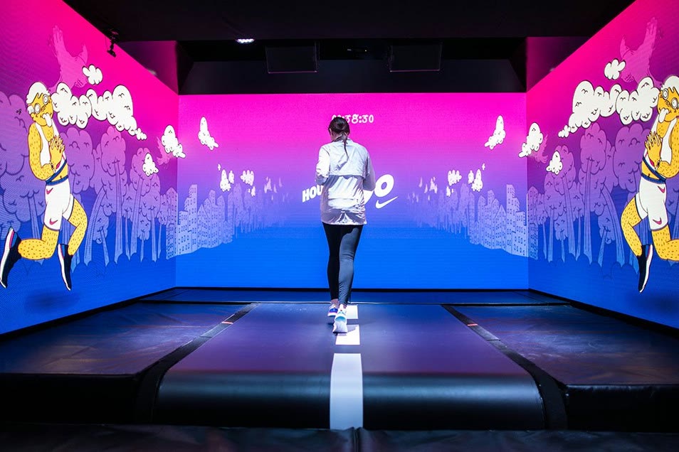 A visitor walking a lit path toward a magenta and blue screen wall, with illustrated figures on the side walls: the Nike House of Go pop-up.