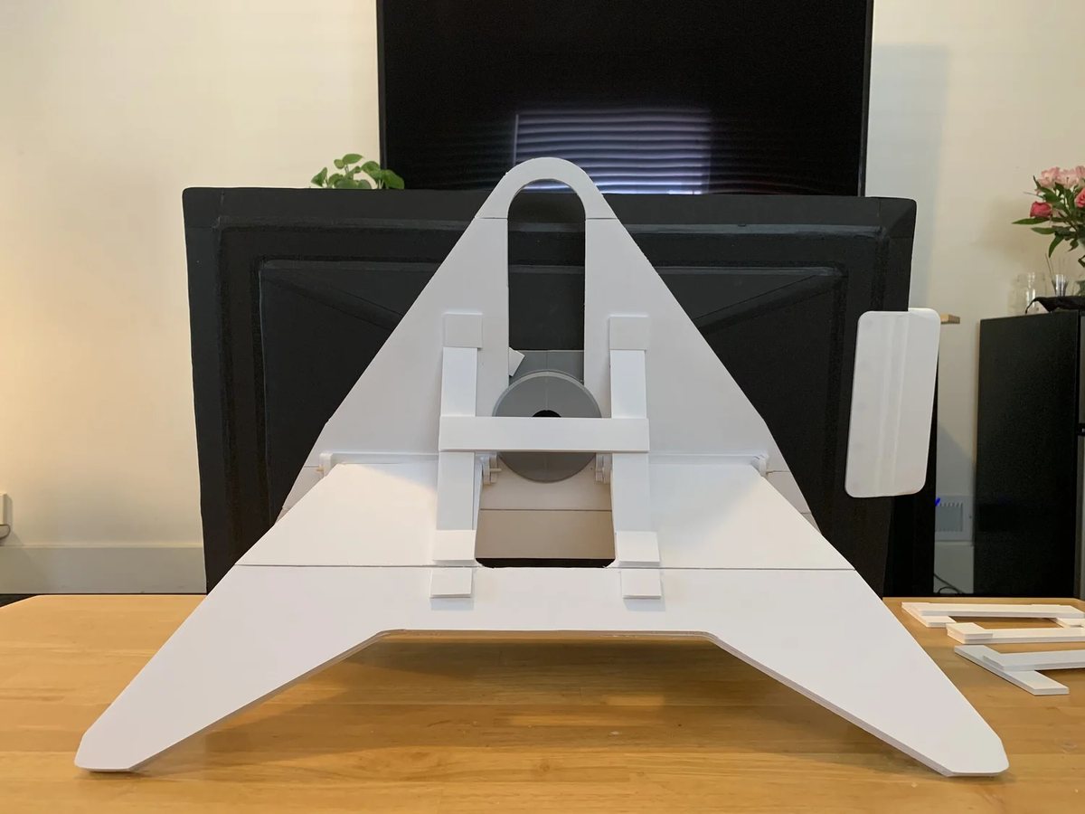 A white 1:1 scale model of a display stand on a desk, propping up a large black display from behind.